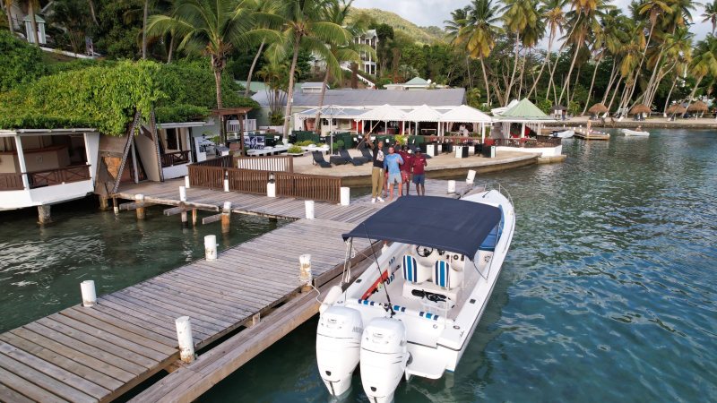 People standing on a dock next to a boat, with tropical huts and palm trees in the background.