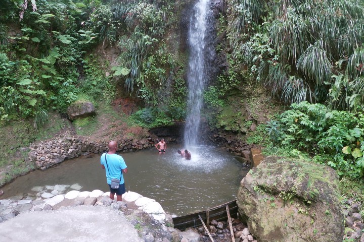 Small waterfall with lush greenery, people swimming, one person standing nearby taking photos.