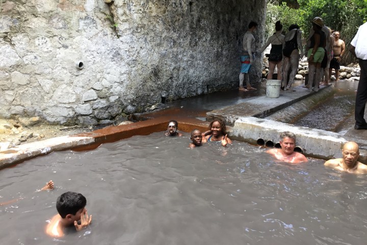 People bathing in a natural hot spring, some covered in mud, near a rocky wall.