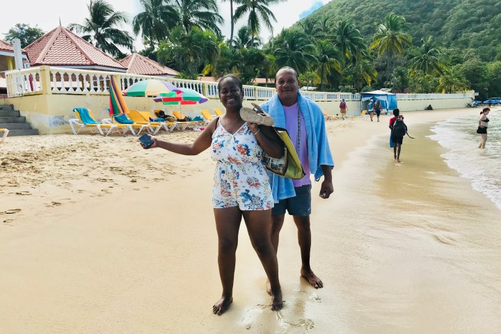 Two people smiling on a sandy beach with colorful umbrellas and palm trees in the background.
