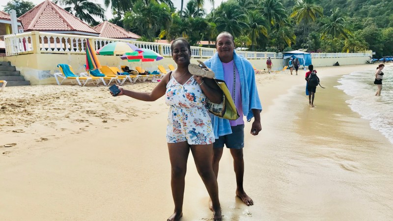 Two people smiling on a sandy beach with colorful umbrellas and palm trees in the background.