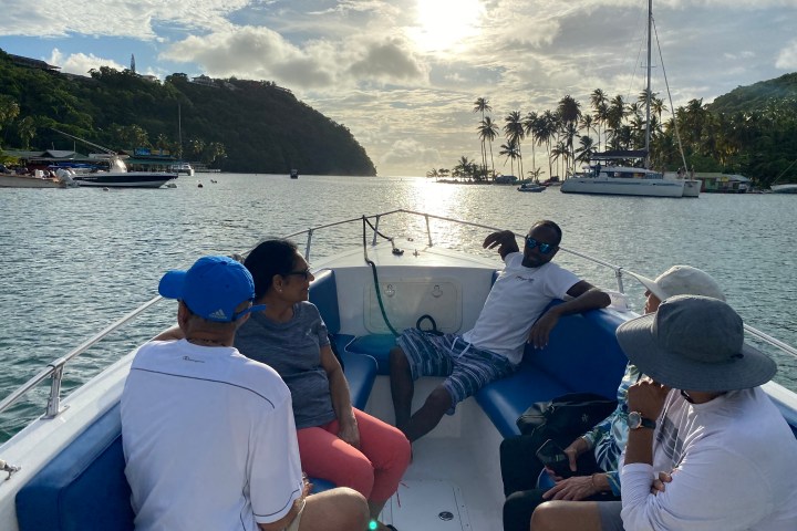Five people relaxed on a boat in a scenic tropical bay with palm trees and sailboats at sunset.