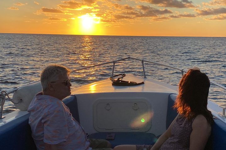 Two people on a boat watching a sunset over the ocean, with scattered clouds in the sky.