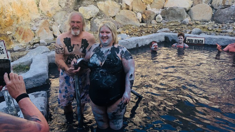 People covered in mud standing in a rocky hot spring with mineral deposits on the walls.