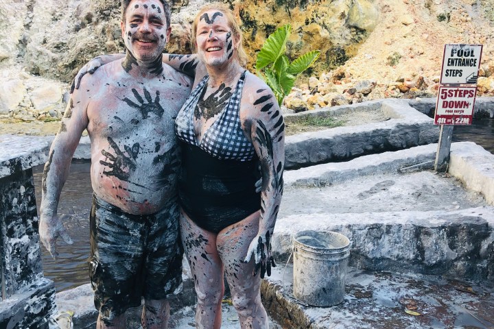 Two people covered in mud smiling with rocky background, sign indicating pool entrance and steps.