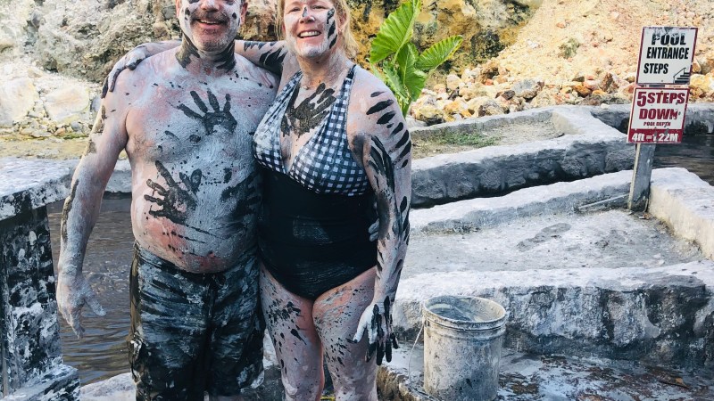 Two people covered in mud smiling with rocky background, sign indicating pool entrance and steps.