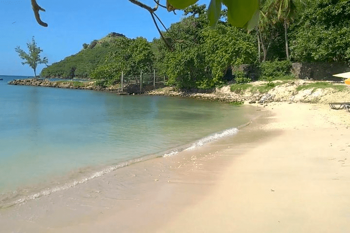 Quiet tropical beach with clear water, trees, and distant hills under a blue sky.