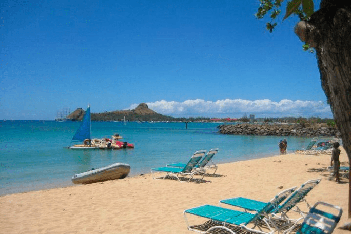 Sunny beach with lounge chairs, sailboat, and distant hills on clear blue water.
